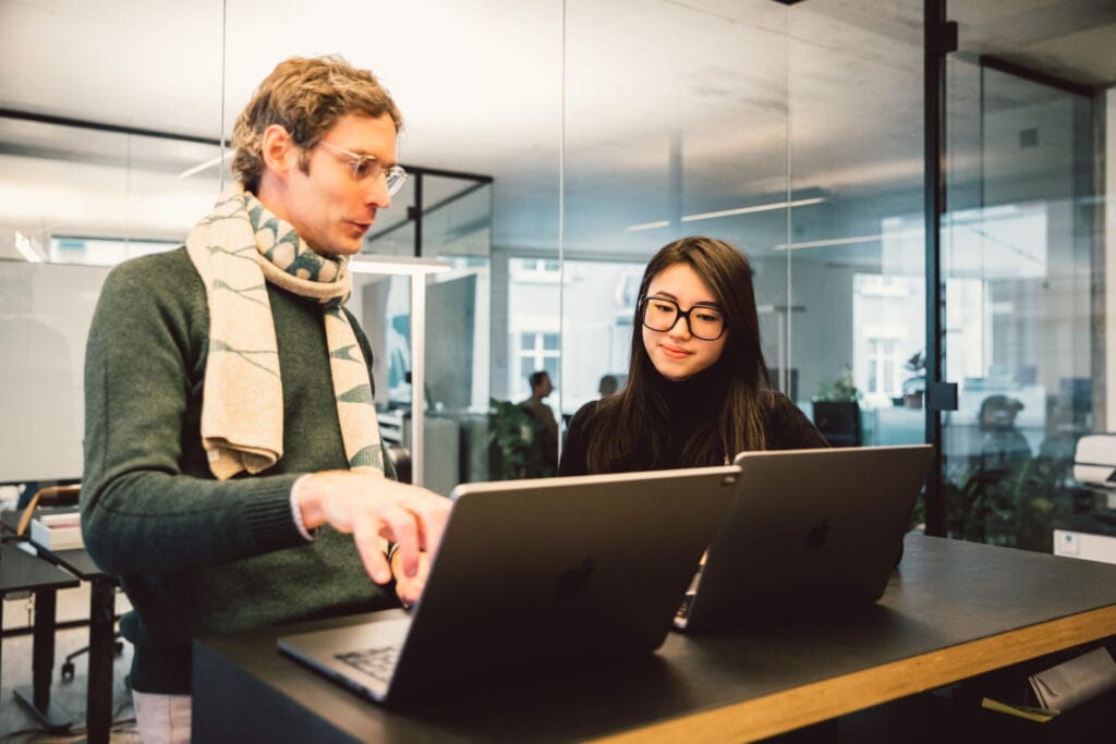 Mann und Frau stehen vor zwei Laptops auf einem Hochtisch und Diskutieren was auf dem Bildschirm ist